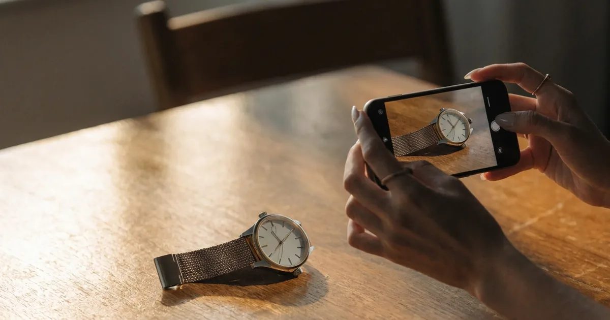 Una mano fotografa con il telefono un orologio da polso appoggiato su un tavolo di legno; l'orologio appare anche sullo schermo del telefono.