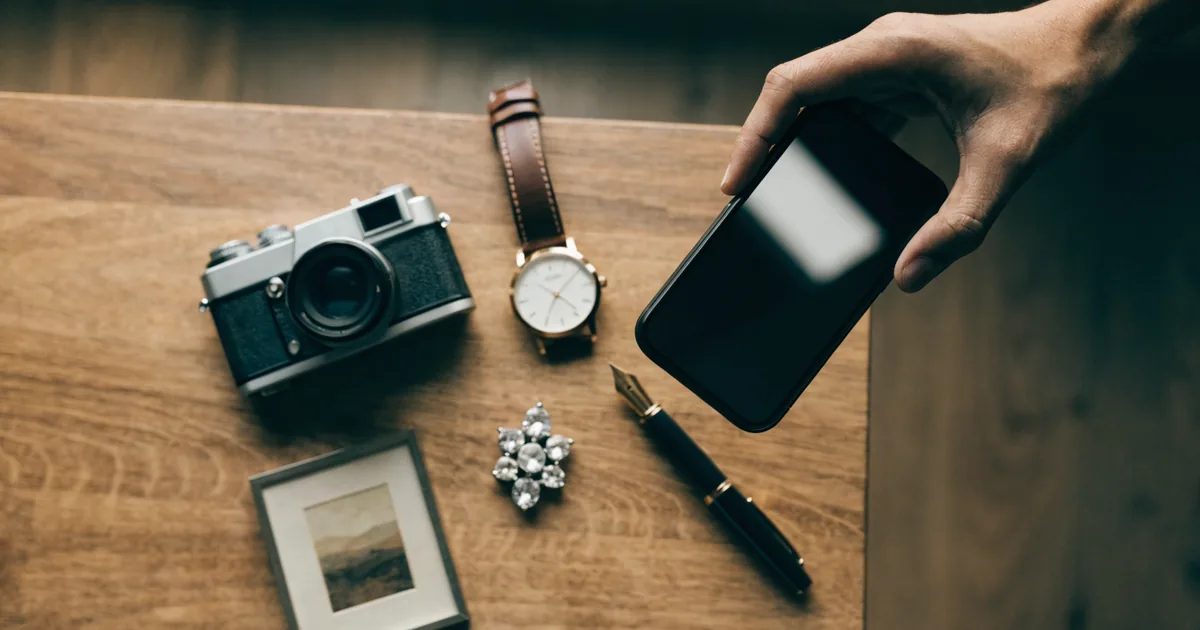 Veduta dall'alto di un tavolo di legno con oggetti di valore — una macchina fotografica classica, un orologio da polso, una spilla di diamanti, una penna stilografica e una piccola cornice — mentre una mano li fotografa con un telefono per documentarli.
